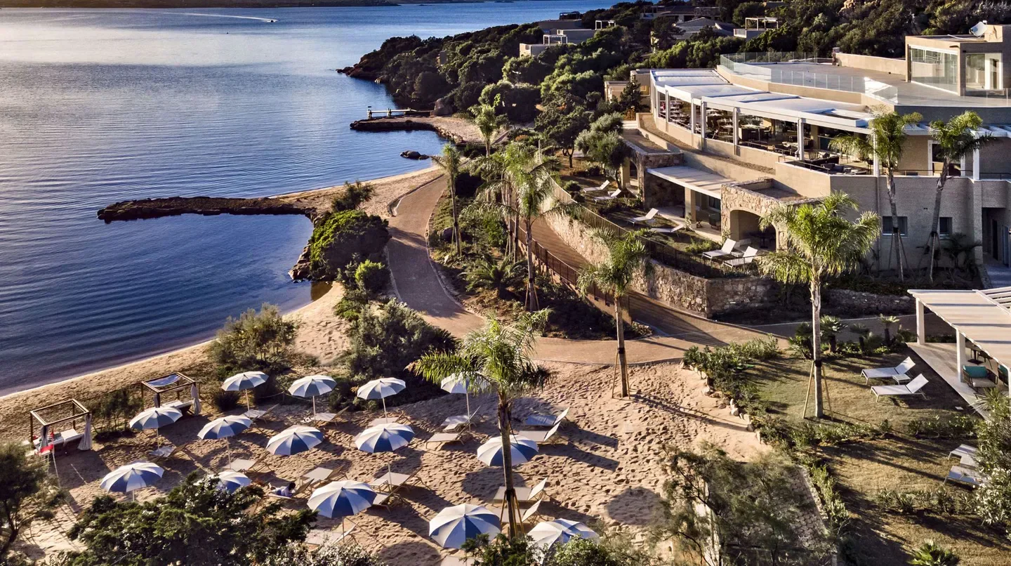 Beachfront view of 7Pines Resort Sardinia, with umbrellas and calm sea in Sardinia.